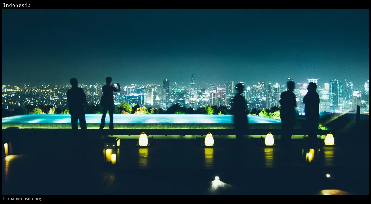 People silhouetted on a terrace with an infinity pool, overlooking a city at night.