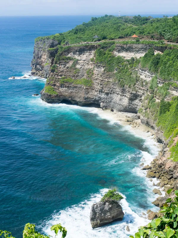 A busy surf beach below a cliff at dusk, warungs and umbrellas built into the rock, a volcano silhouetted across the water.