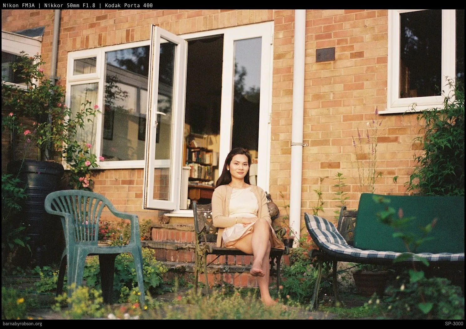 Woman sitting barefoot on garden bench outside brick house.
