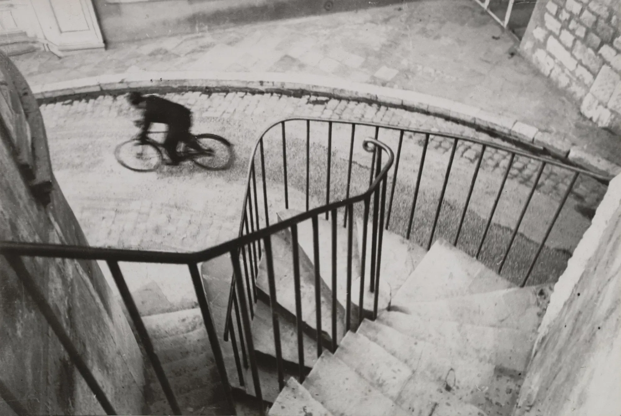A cyclist blurring past below a spiral staircase railing, black and white.