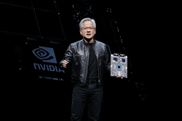 Jensen Huang stands on a dark stage holding an Nvidia accelerator board, with the Nvidia mark projected behind him.