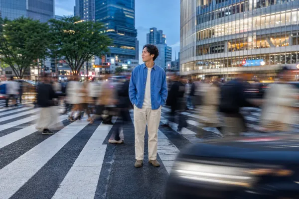 A man in a blue shirt stands still at Shibuya Crossing while crowds and traffic streak past in motion blur.