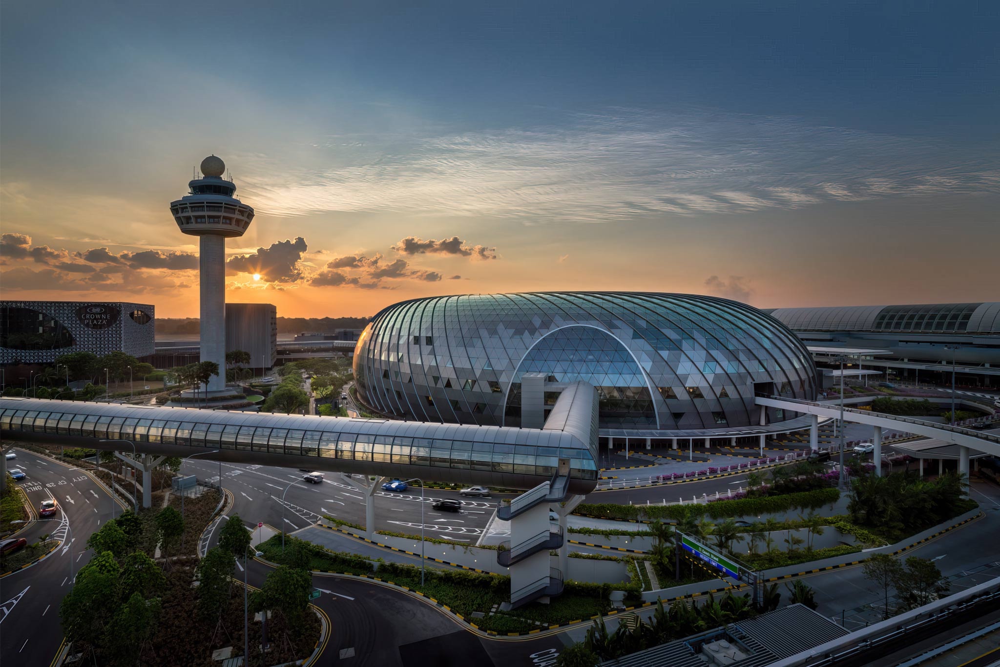 Jewel Changi Airport's glass torus beside the control tower at sunset, landscaped roads running beneath